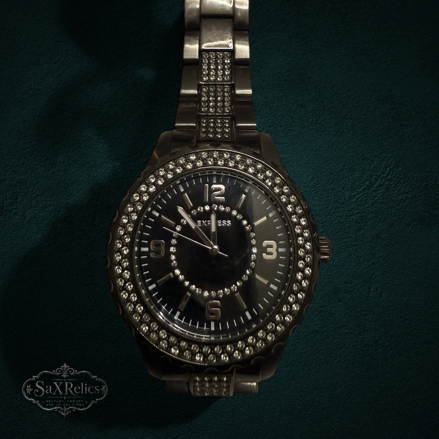 My Store Express dark metal link wristwatch with a crystal-encrusted bezel and black dial on a dark green background.