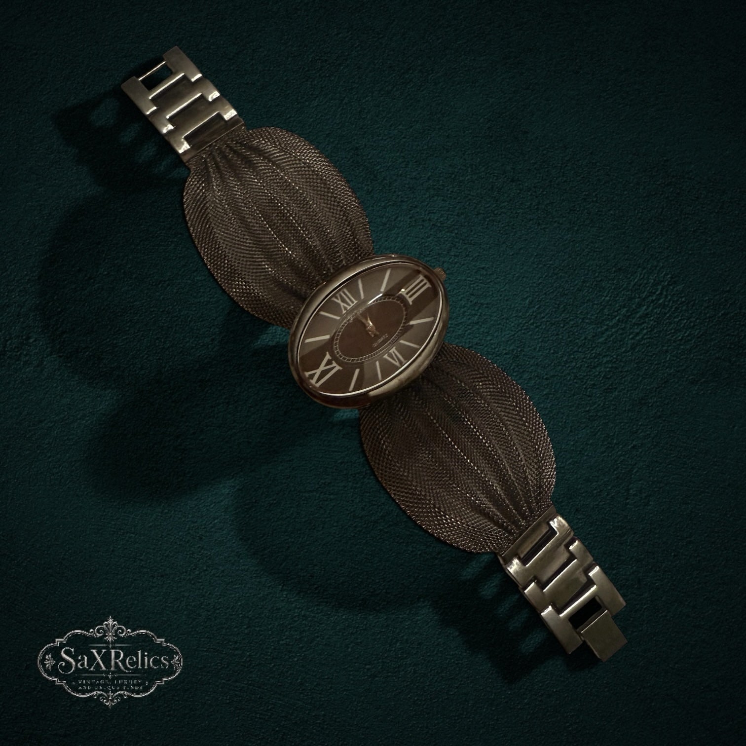My Store Geneva quartz wristwatch with a dark metallic oval dial, Roman numerals, and a textured fan-like mesh bracelet.