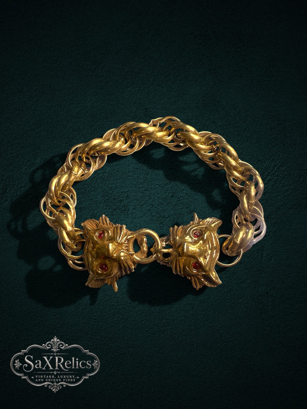 SaX Relics gold-tone link bracelet featuring dual lion heads with red gemstone eyes on a dark green background.